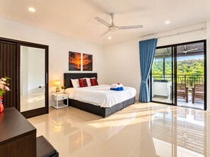 A modern hotel room with a large bed, a ceiling fan, a nightstand with a lamp, a view of greenery through a sliding glass door, and a painting on the wall. - Luxurious Villa With Private Pool and Stunning Views (Chalong)
