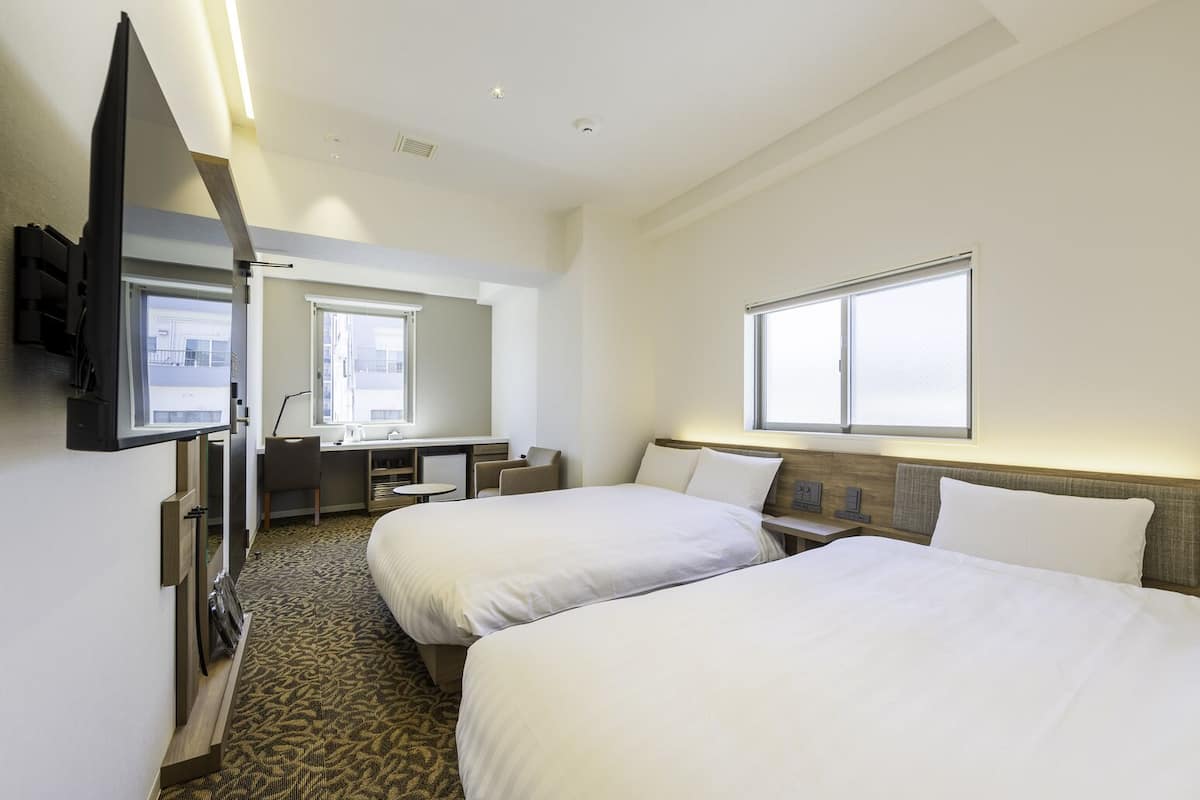 A hotel room with two beds, a desk, a chair, a TV, and a window.