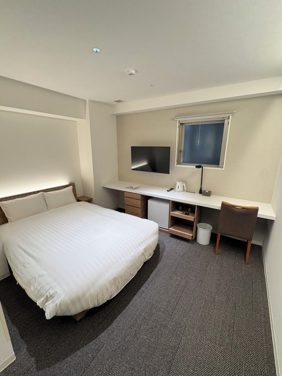 Habitación de hotel con cama, escritorio con televisor, ventana y un pequeño cubo de basura.