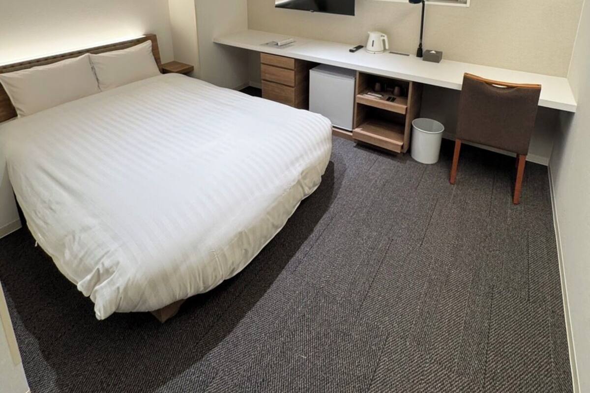A hotel room with a bed, a desk with a TV, a window, and a small trash bin.