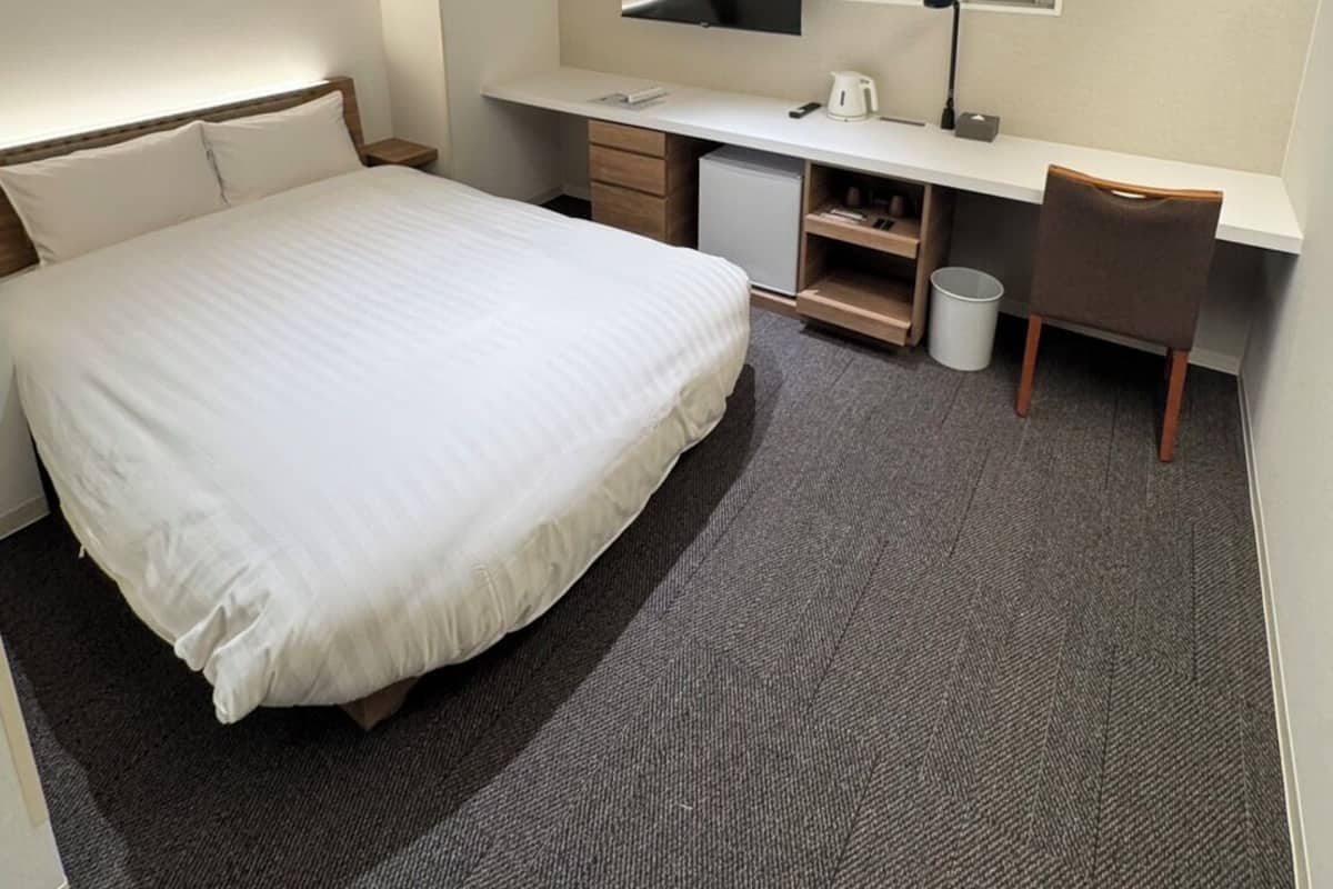 A hotel room with a bed, a desk with a TV, a window, and a small trash bin.