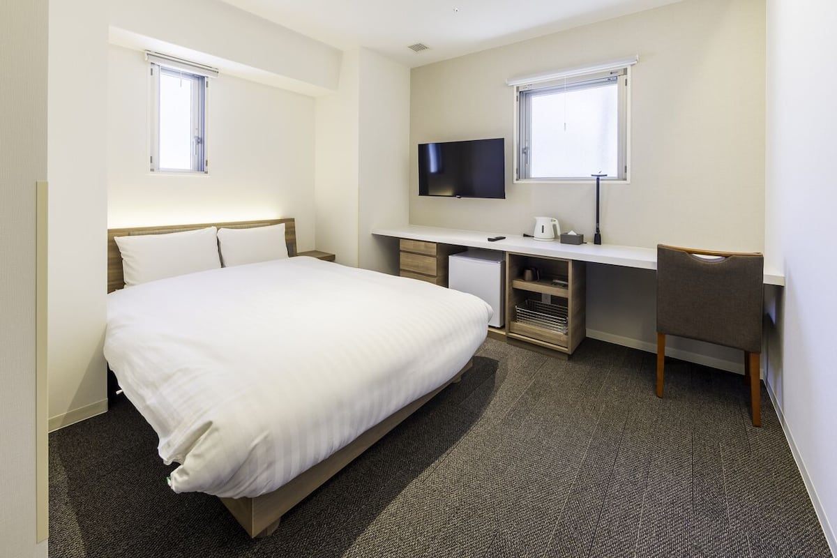 A hotel room with a bed, desk, chair, TV, and two windows.