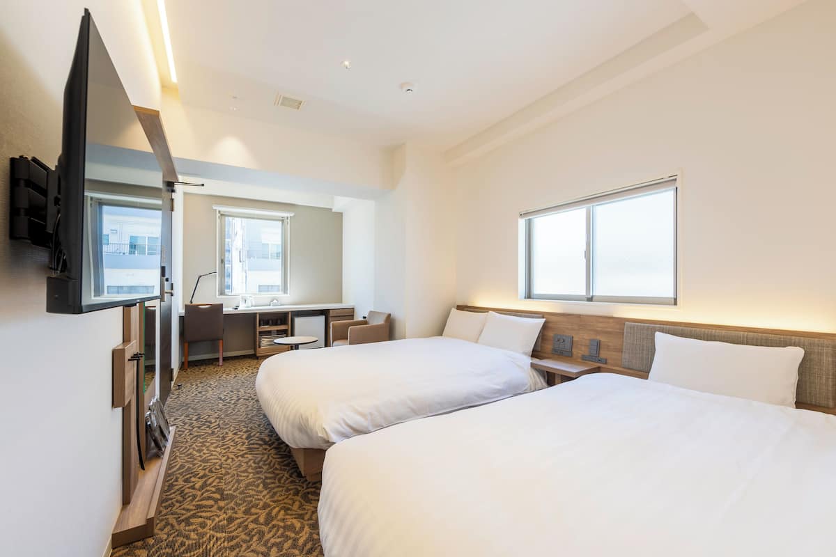 A hotel room with two beds, a desk, a chair, a TV, and a window.