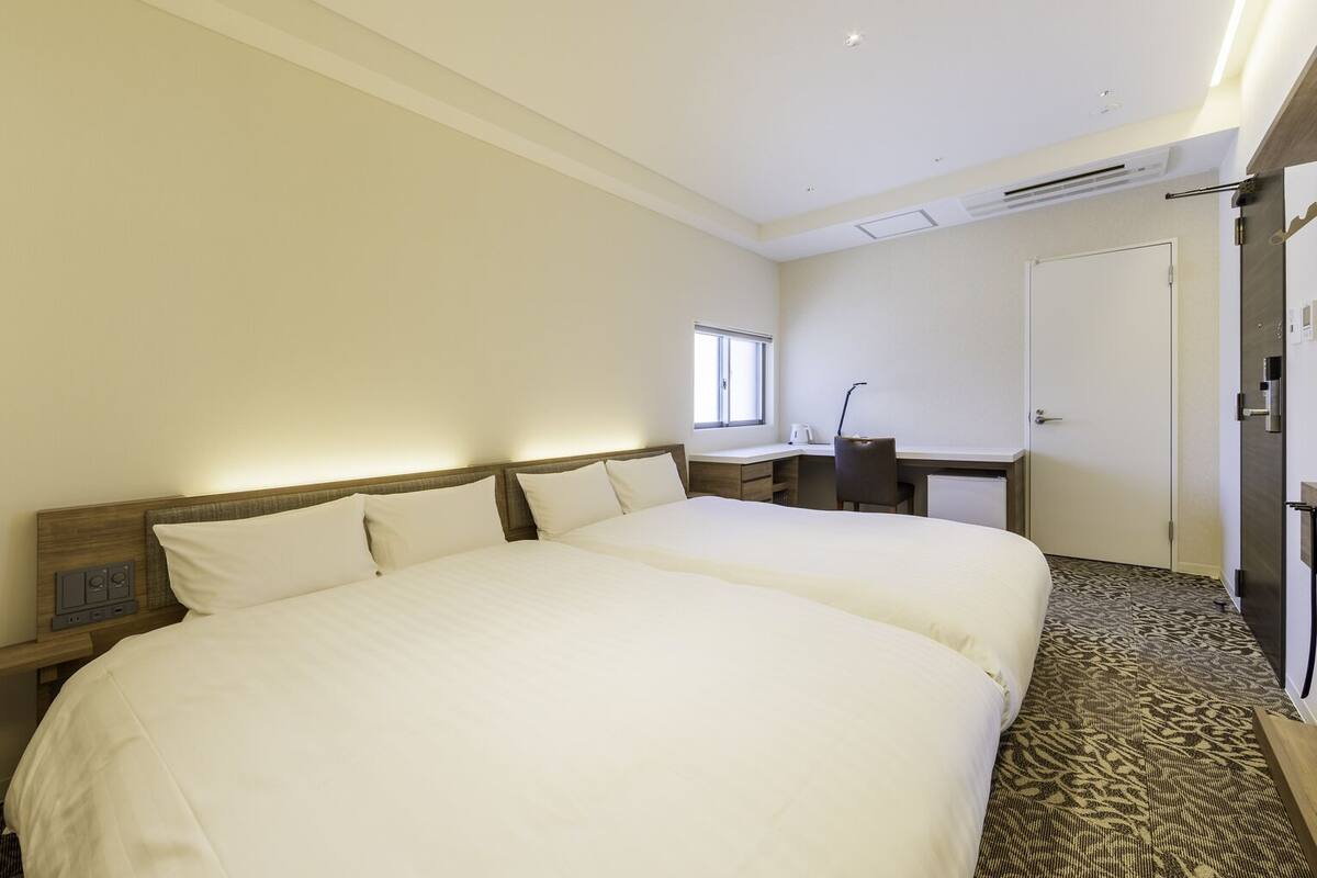 A hotel room with two beds, a desk, a chair, a wardrobe, and a window.