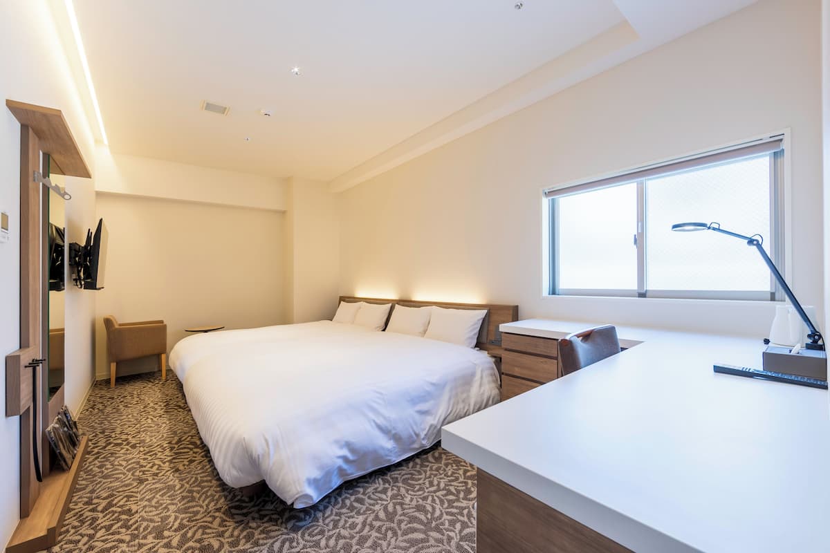 A modern hotel room with a large bed, bedside table, desk, and a window.