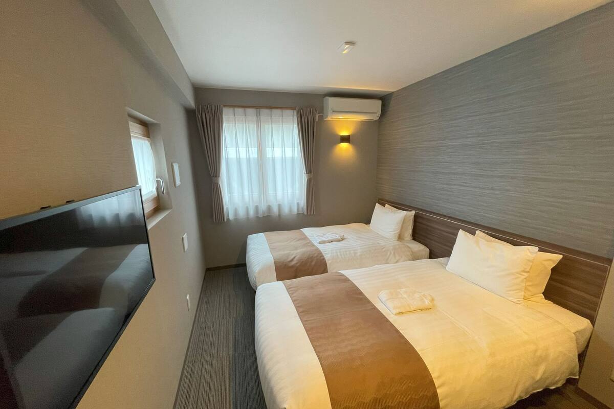 A hotel room with two beds, a flat-screen TV, an air conditioning unit, and a window with curtains.