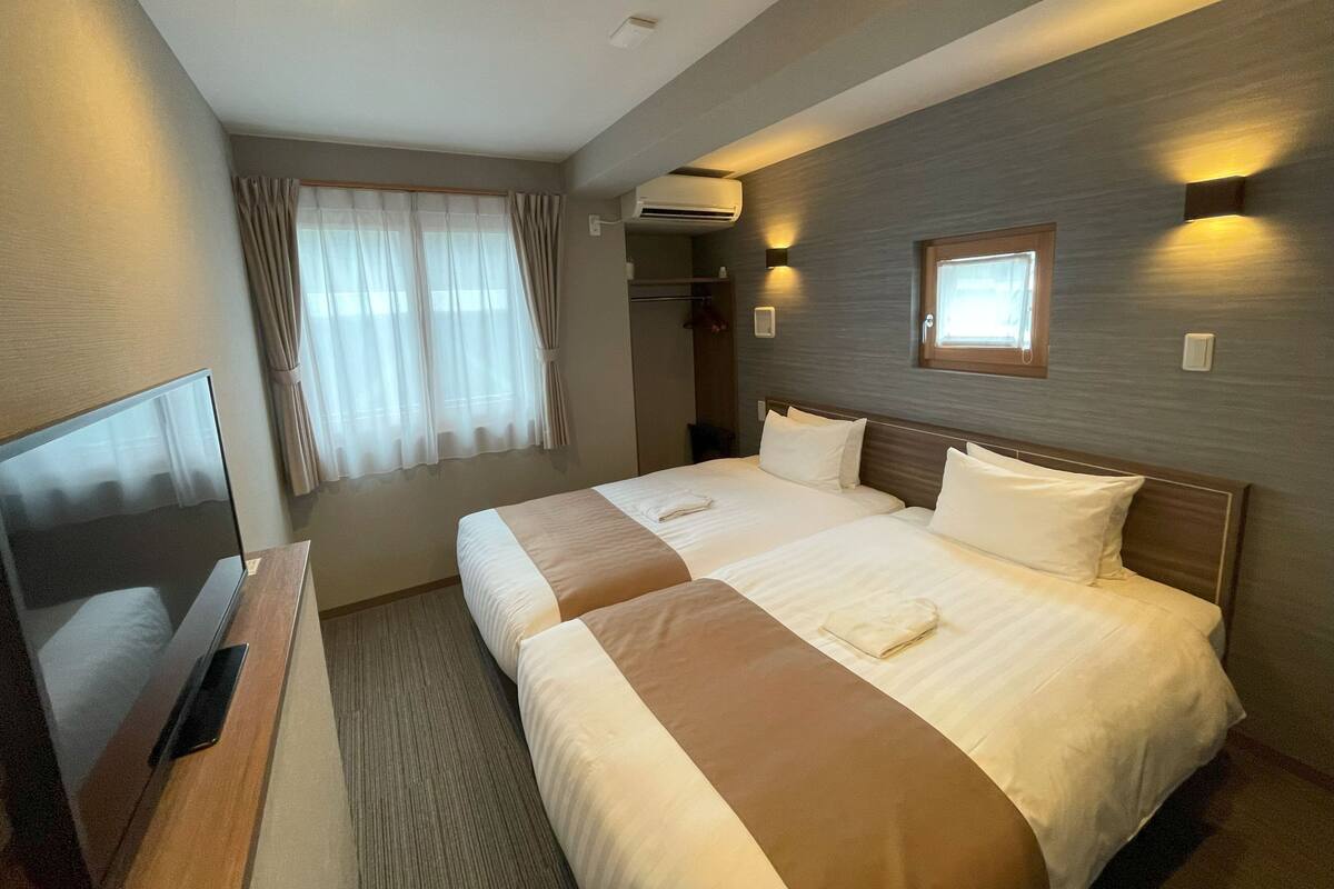 A hotel room with two beds, a TV, an air conditioning unit, and a window with curtains.