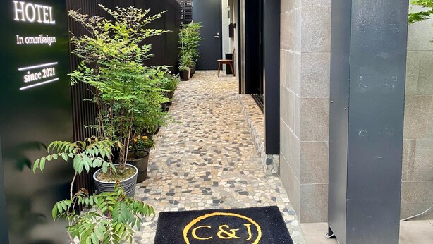 L’entrée d’un hôtel, avec un chemin dallé, des plantes en pot et un paillasson noir orné du logo de l’hôtel.