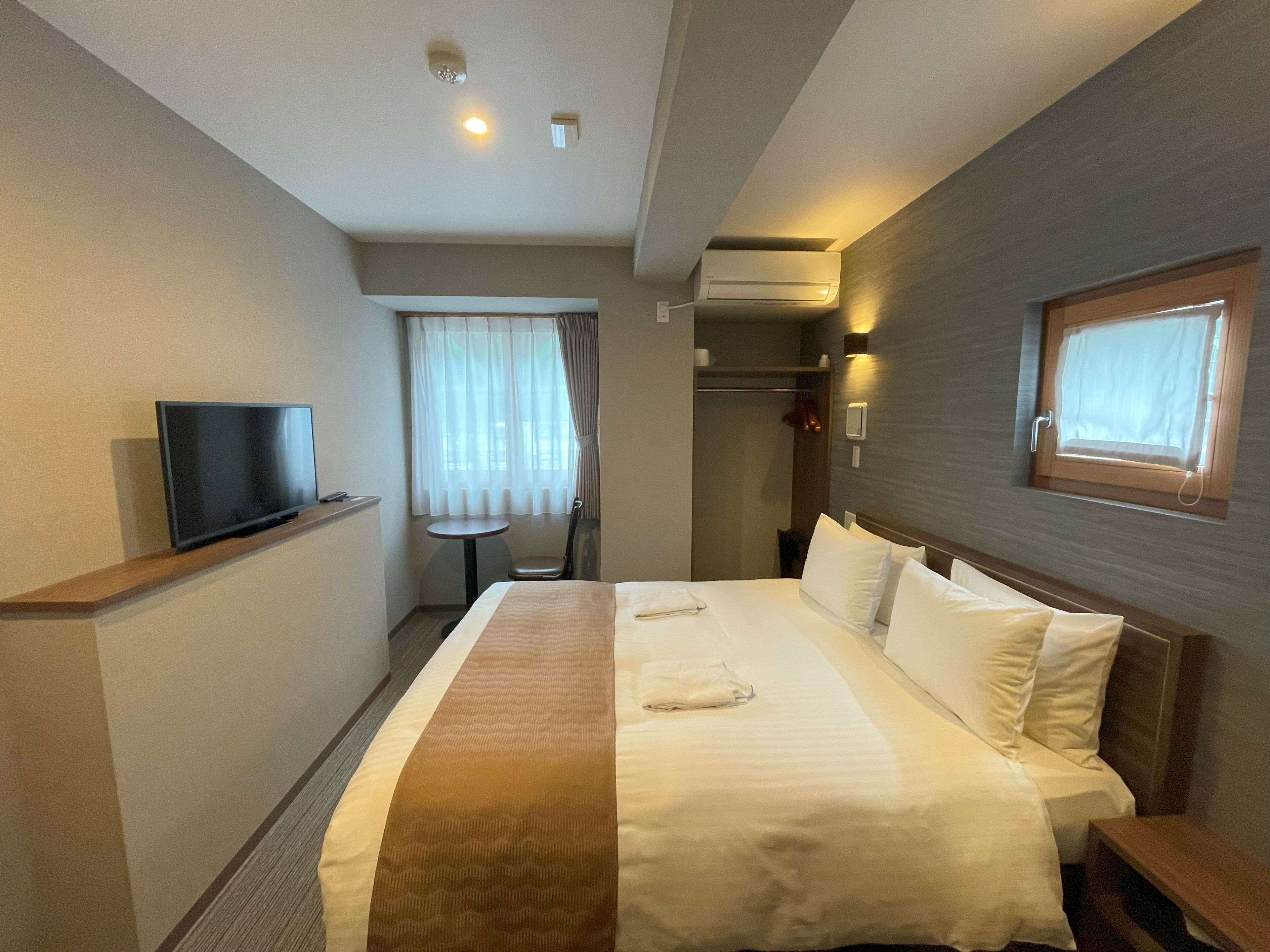 Ein Hotelzimmer mit Bett, Fernseher, einem kleinen Tisch, einem Fenster und einer Klimaanlage.