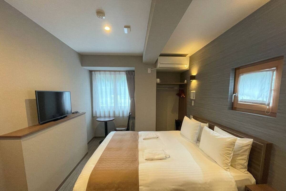 A hotel room with a bed, a TV, a small table, a window, and an air conditioning unit.