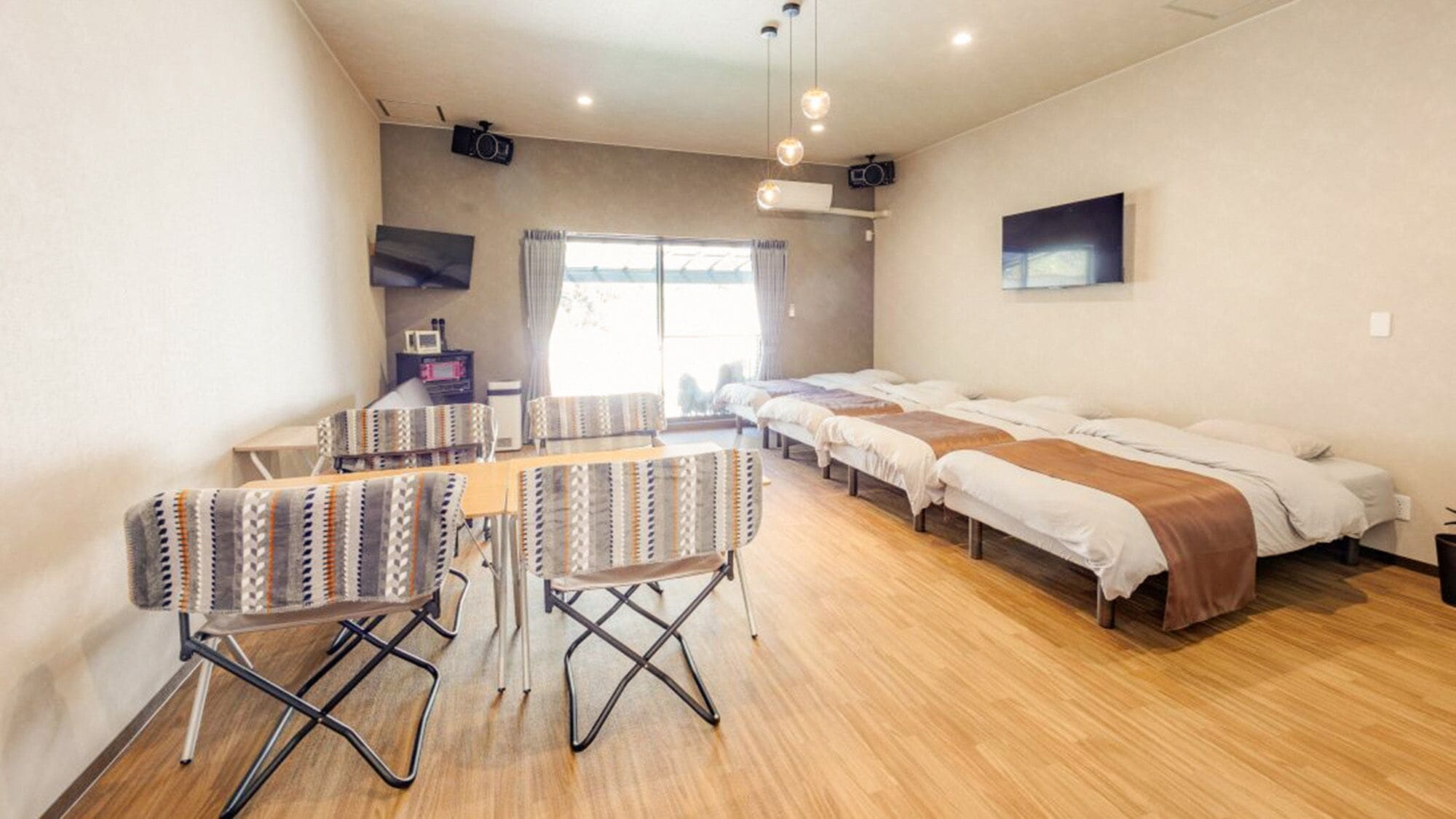 A hotel room with a long bed, a small table with chairs, a TV, and a projector on the ceiling.
