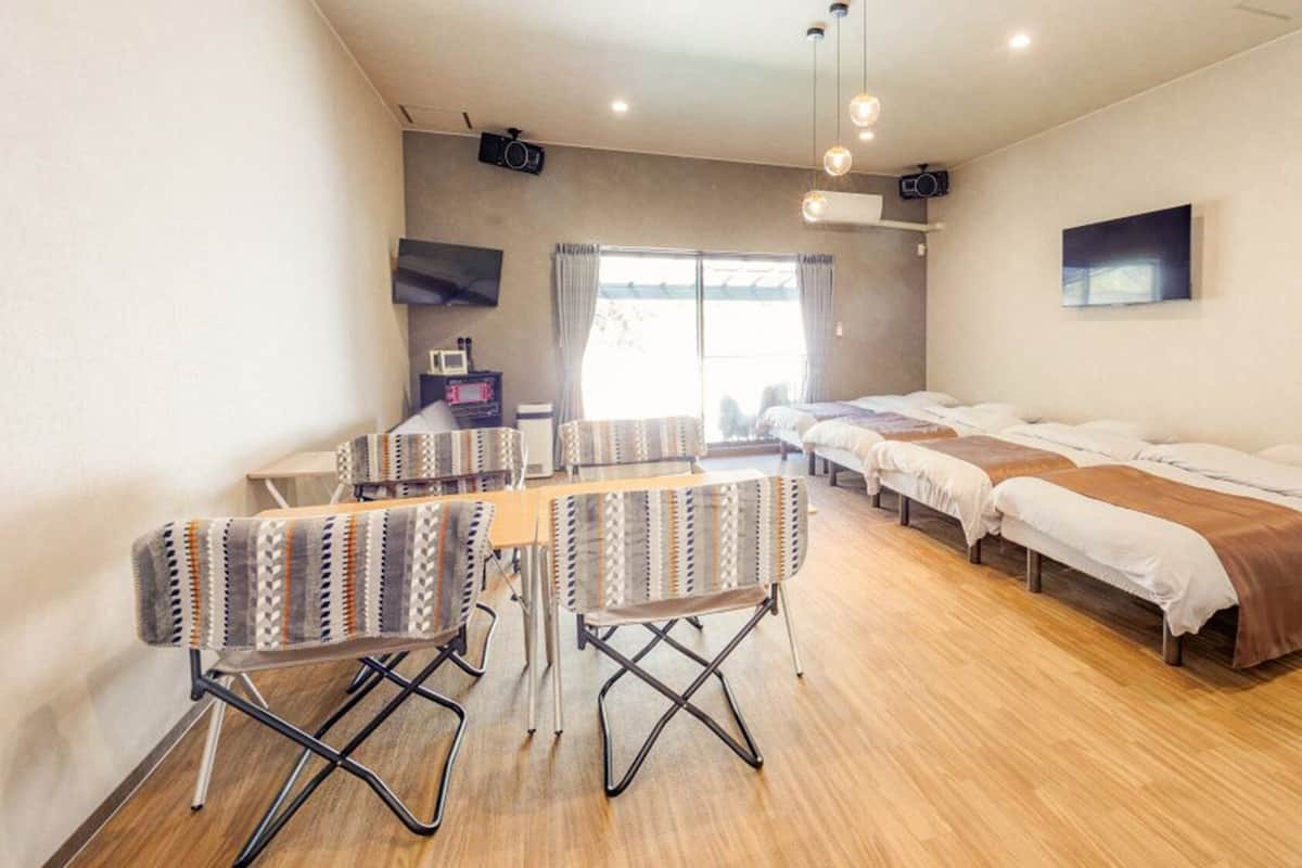 A hotel room with a long bed, a small table with chairs, a TV, and a projector on the ceiling.