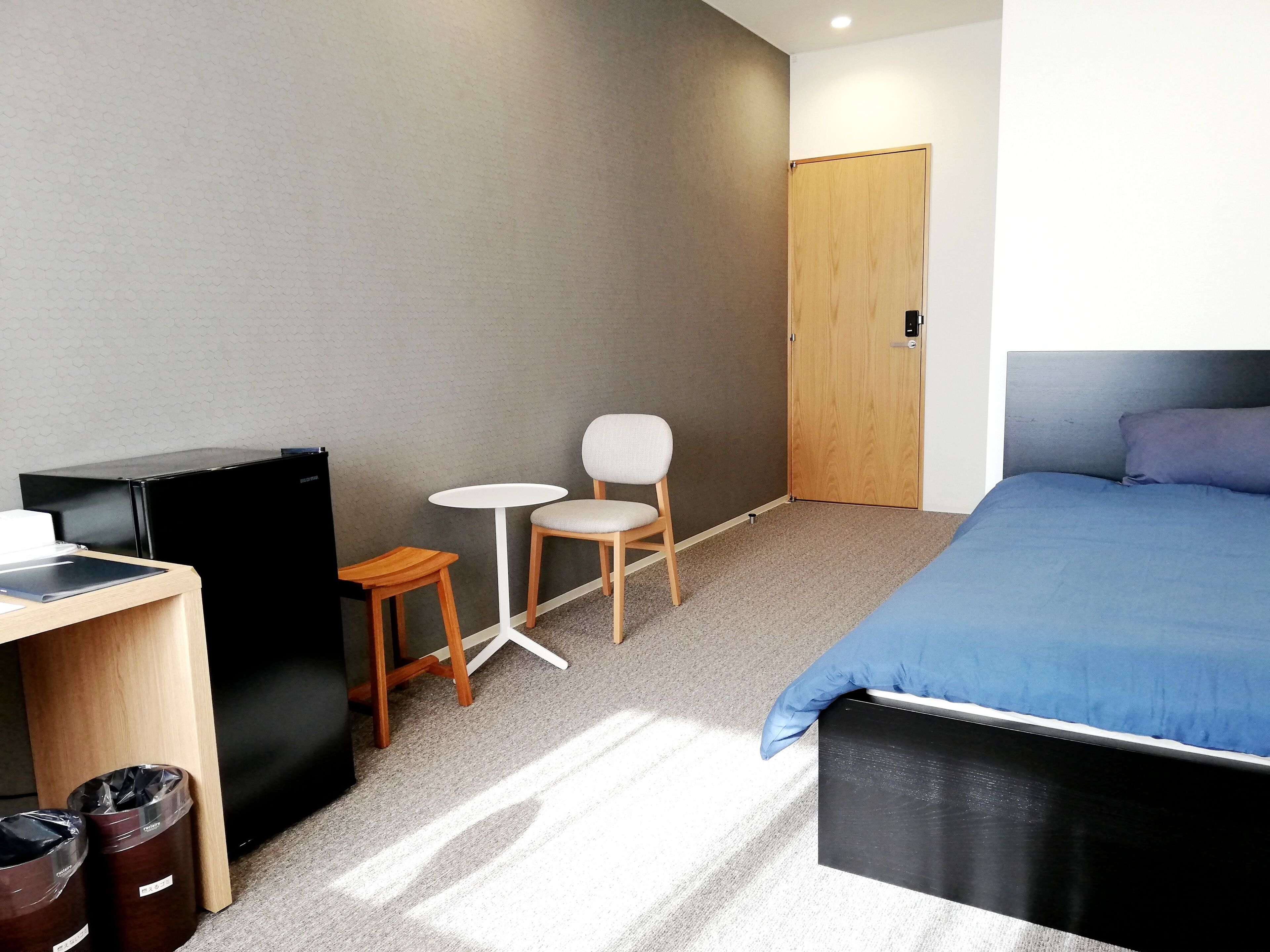 Une chambre à coucher comprenant un lit, un bureau, une chaise, une petite table, une armoire et deux poubelles.
