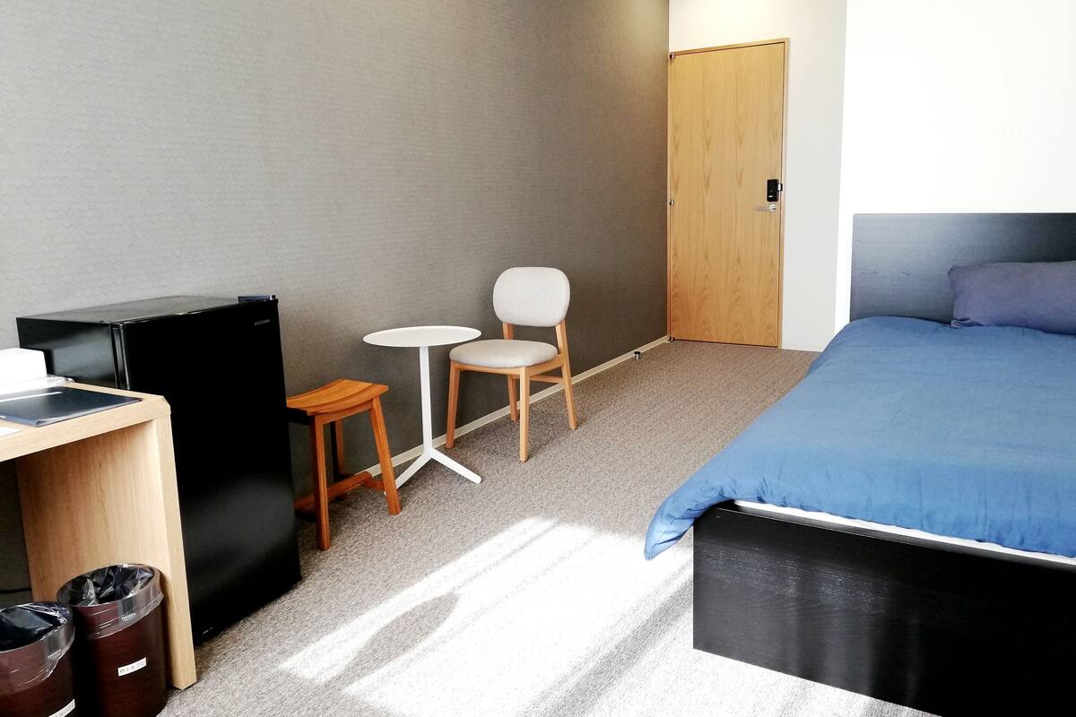 Ein Schlafzimmer mit einem Bett, einem Schreibtisch, einem Sessel, einem kleinen Tisch, einem Kleiderschrank und zwei Mülleimern.