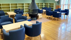 Et hyggeligt opholdsområde indendørs med blå stole og borde, en central sort stov og grønne reoler i baggrunden.