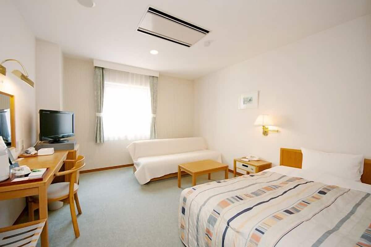 A hotel room with a bed, a desk with a television, a chair, a bedside table, and a window with curtains.