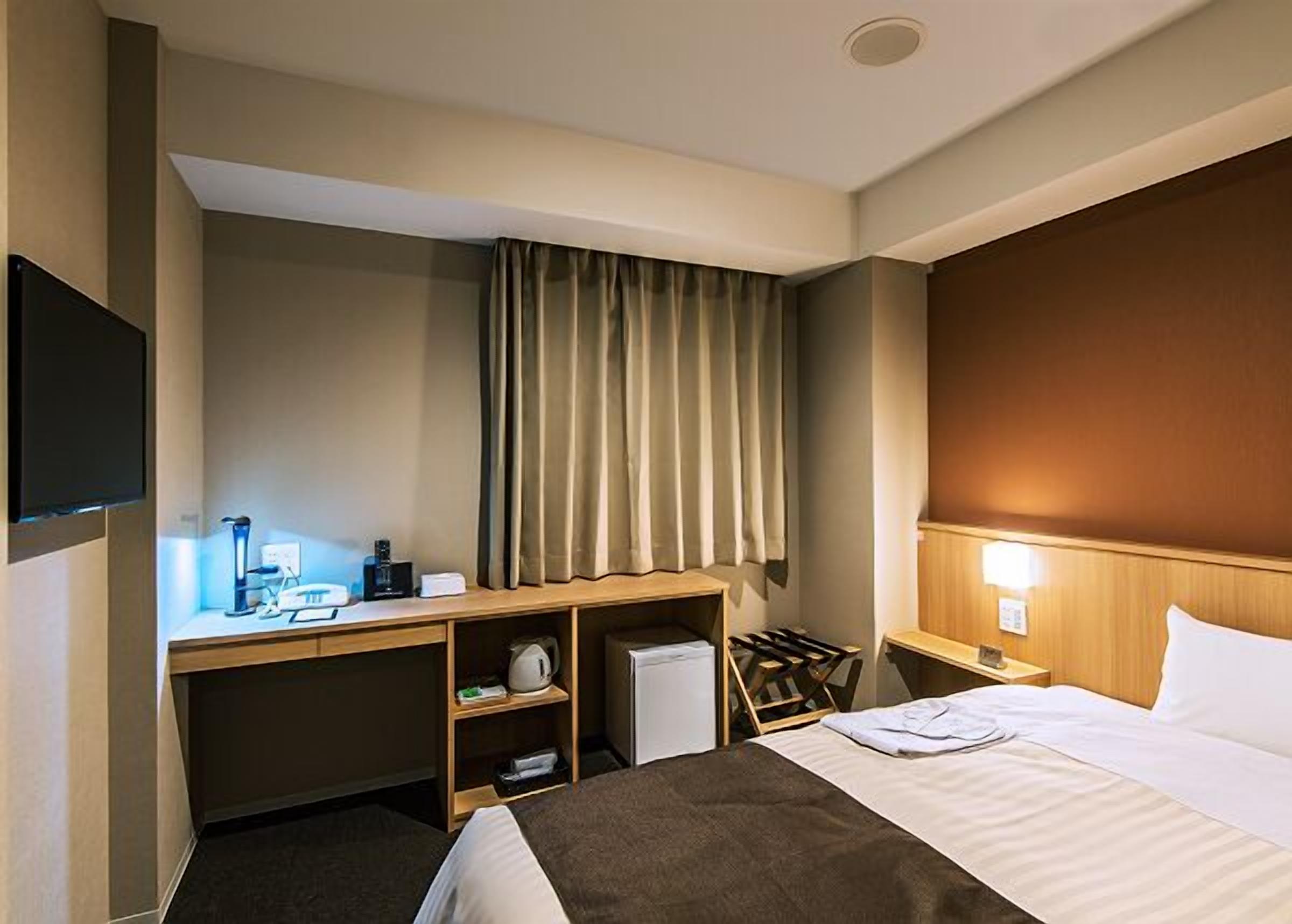 A hotel room with a bed, a desk with a computer, a chair, a TV, and a window with curtains.