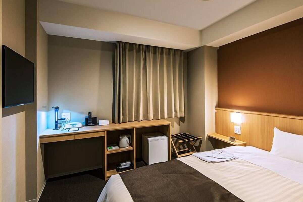 A hotel room with a bed, a desk with a computer, a chair, a TV, and a window with curtains.