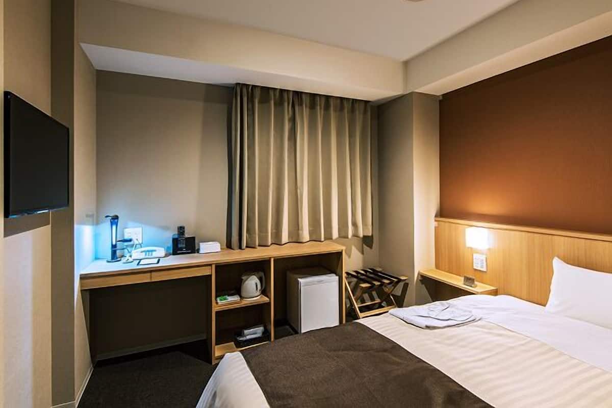 A hotel room with a bed, a desk with a computer, a chair, a TV, and a window with curtains.