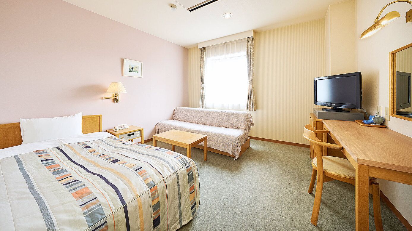 Habitación de hotel con cama, escritorio, televisor, lámpara y ventana con cortinas.