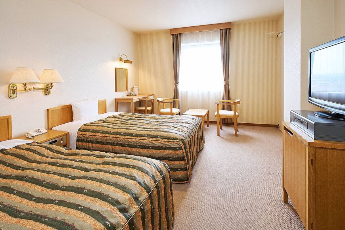A hotel room with two beds, a desk, a chair, a television, and a window with curtains.