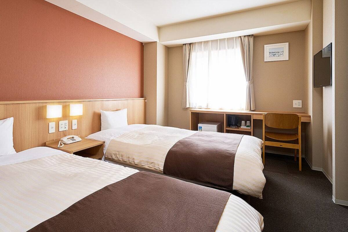 A hotel room with two beds, a desk, a chair, a TV, and a window with curtains.