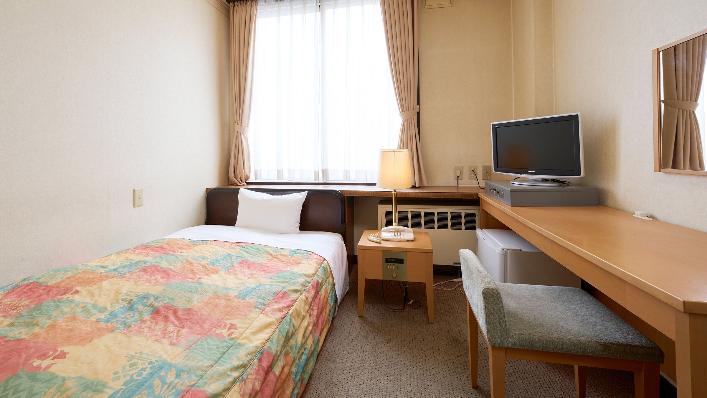 A hotel room with a bed, a desk with a television, a chair, and a small table with a lamp.