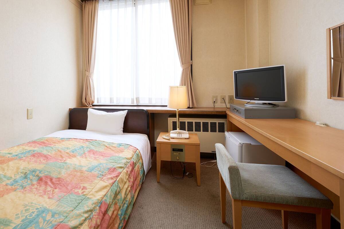 A hotel room with a bed, a desk with a television, a chair, and a small table with a lamp.