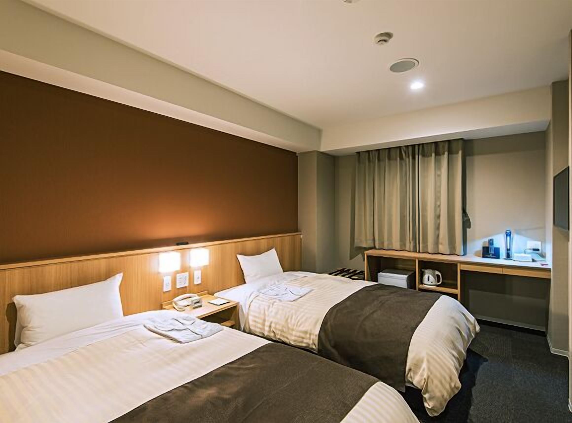A hotel room with two beds, a desk, a chair, a small table, and a window with curtains.