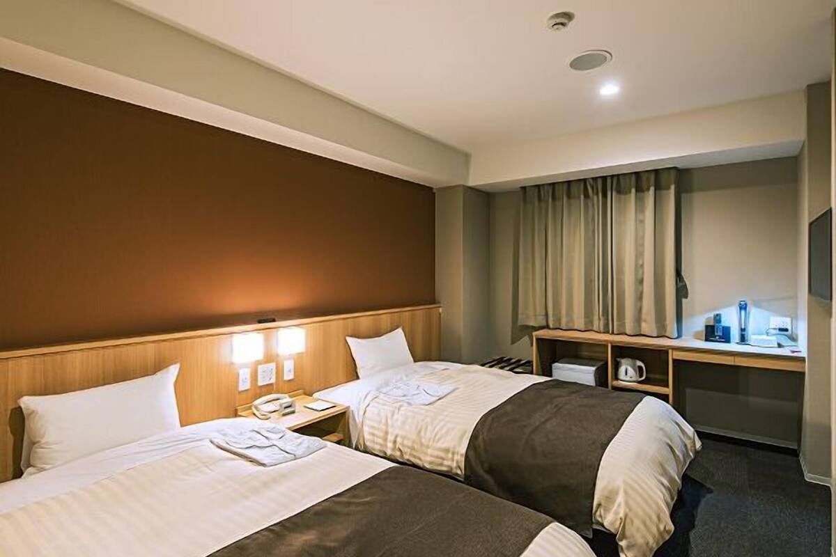 A hotel room with two beds, a desk, a chair, a small table, and a window with curtains.