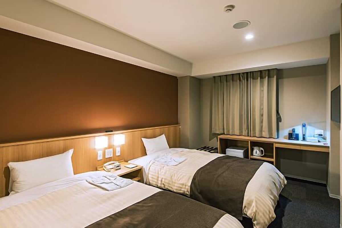 A hotel room with two beds, a desk, a chair, a small table, and a window with curtains.