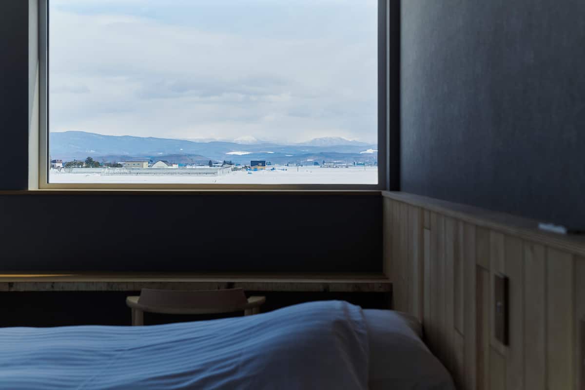 Una habitación con una cama, una silla y un ventanal con vistas a un paisaje nevado y montañas.