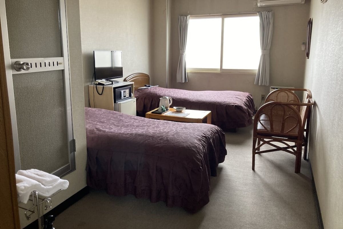 Habitación de hotel con dos camas, un escritorio con computadora, una silla y un ventanal con cortinas.