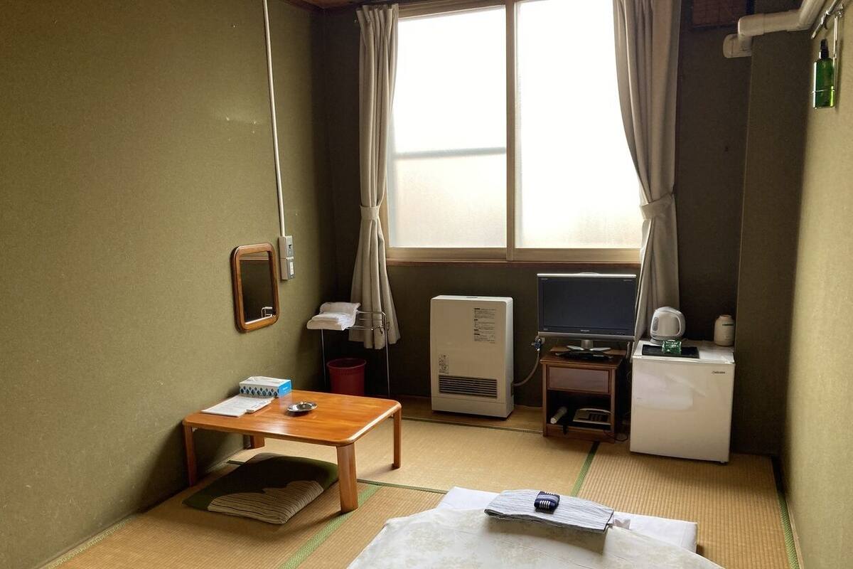 Hotellihuone, jossa on sänky, pieni pöytä, työpöytä, televisio, lämmitin ja ikkuna, jossa on verhot.
