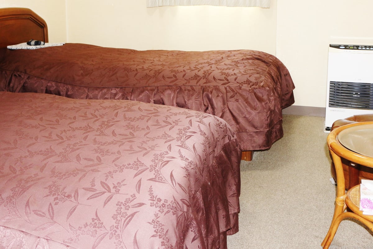 Una habitación con dos camas, una silla, un calefactor y una ventana con cortinas translúcidas.