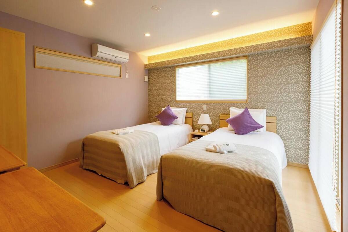 A hotel room with two beds, a wooden table, an air conditioning unit, and a window with blinds.