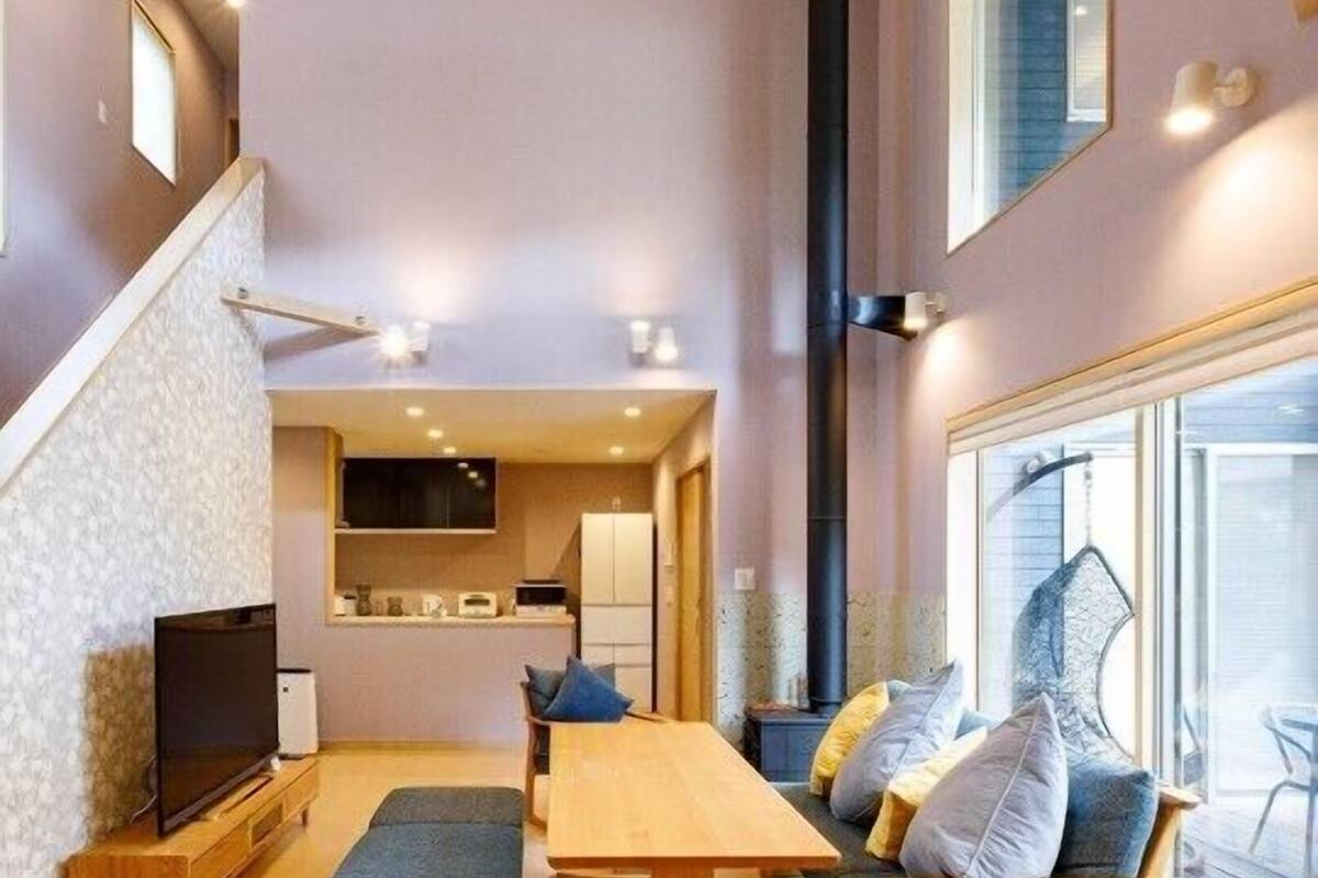 A modern living room with a wooden table, a bench, and a sofa with cushions. There is a TV on a stand, a kitchen area in the background, and a large window with a view.