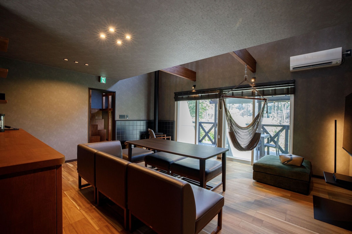 A modern interior with a wooden dining table, benches, a hanging chair, and a large window with a view.