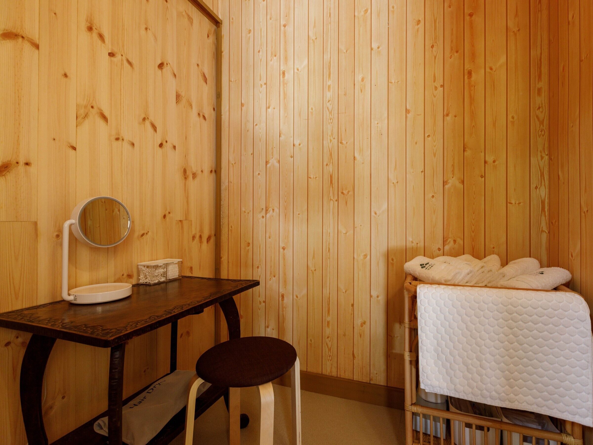 Een houten kamer met een houten tafel, een kruk, een bed met een witte sprei en een ronde spiegel aan de muur.