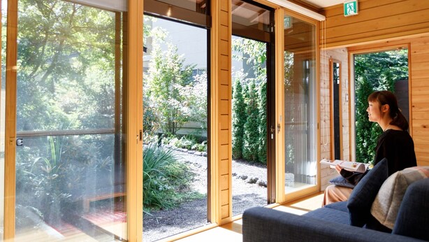 A room with large sliding glass doors leading to a garden, a sofa, and a person reading a book. - Morihaku (Karuizawa)