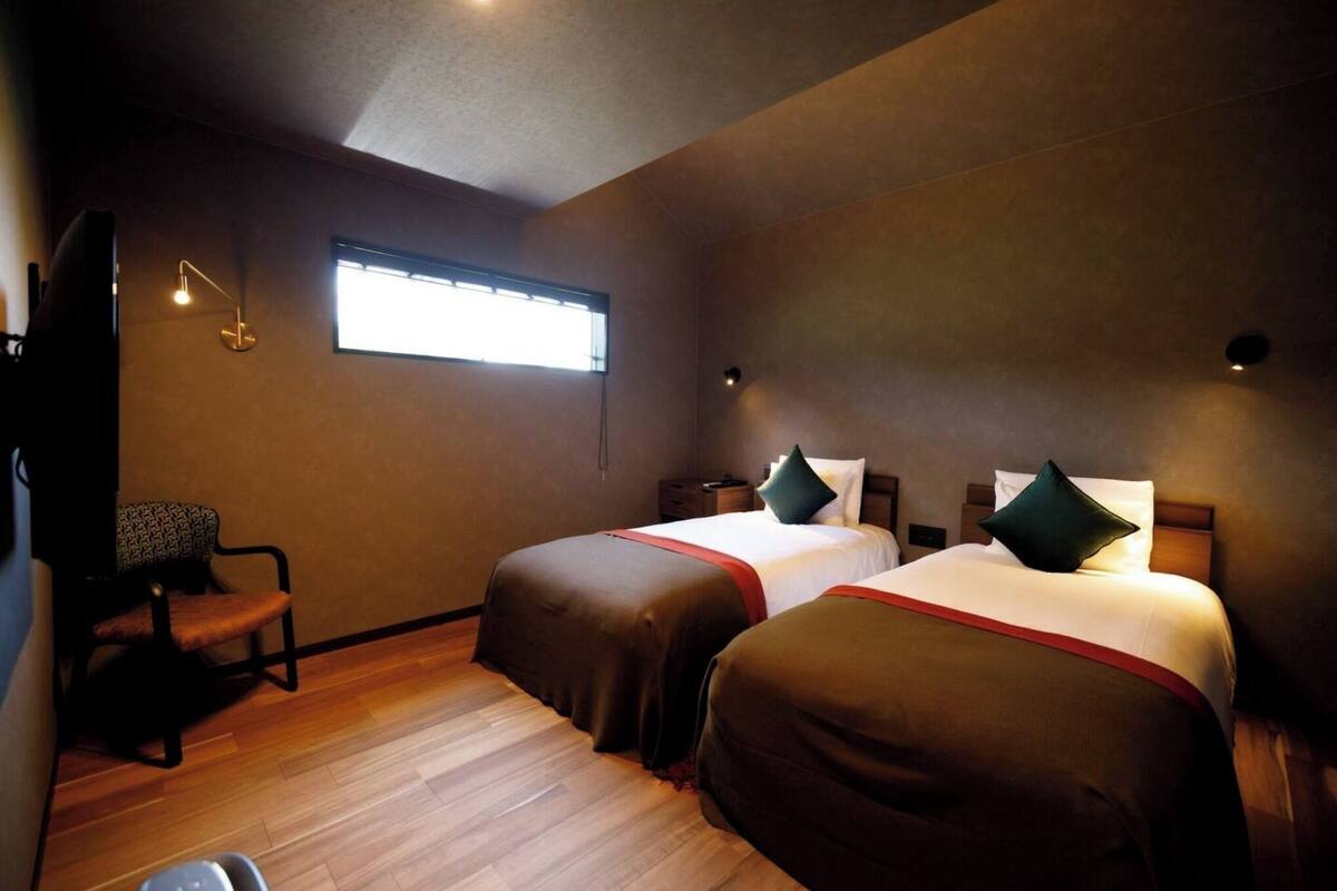 A hotel room with two beds, a chair, a desk, and a window.