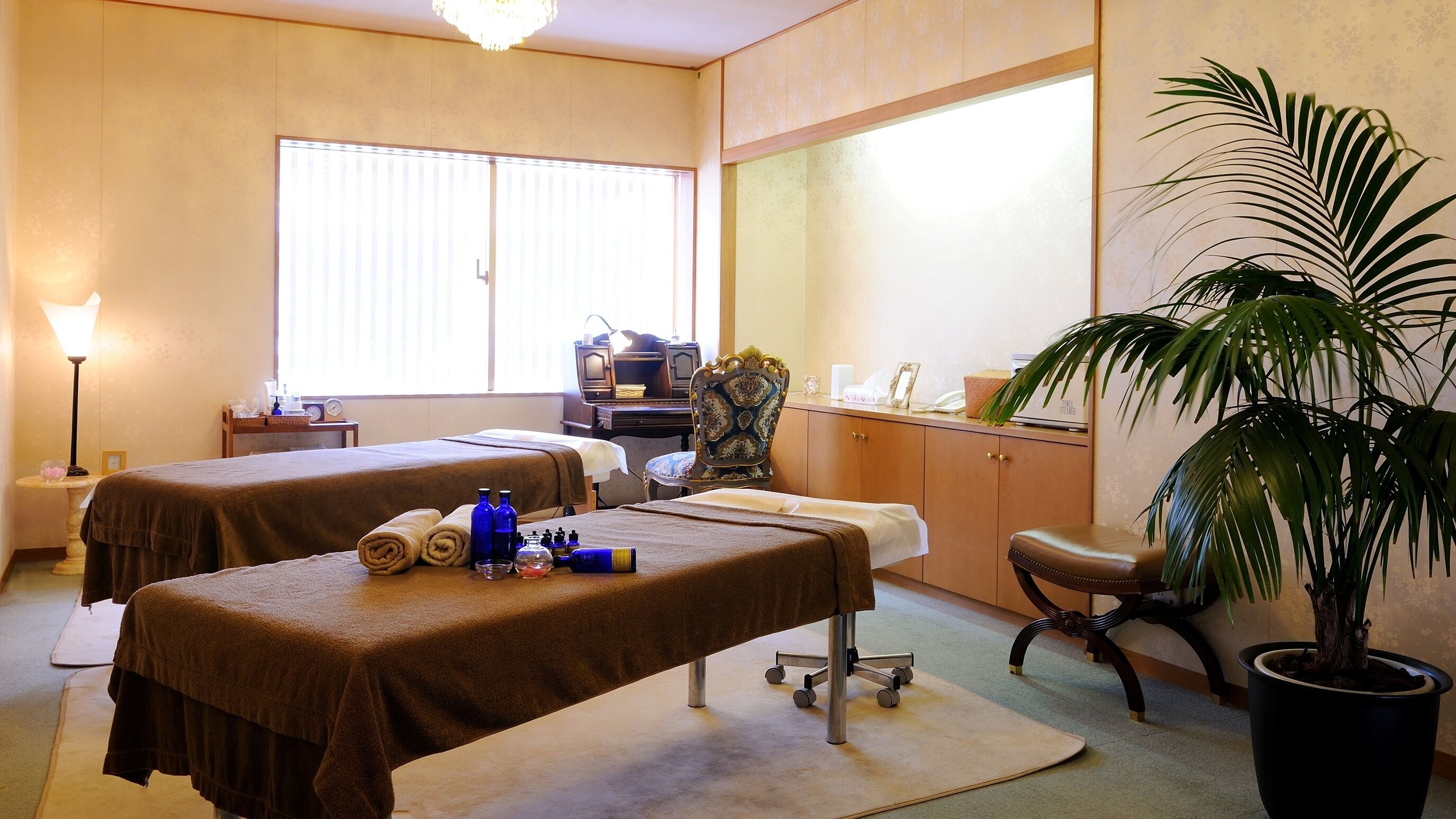 A room with two massage tables, a desk, a chair, a large mirror, and a potted plant.