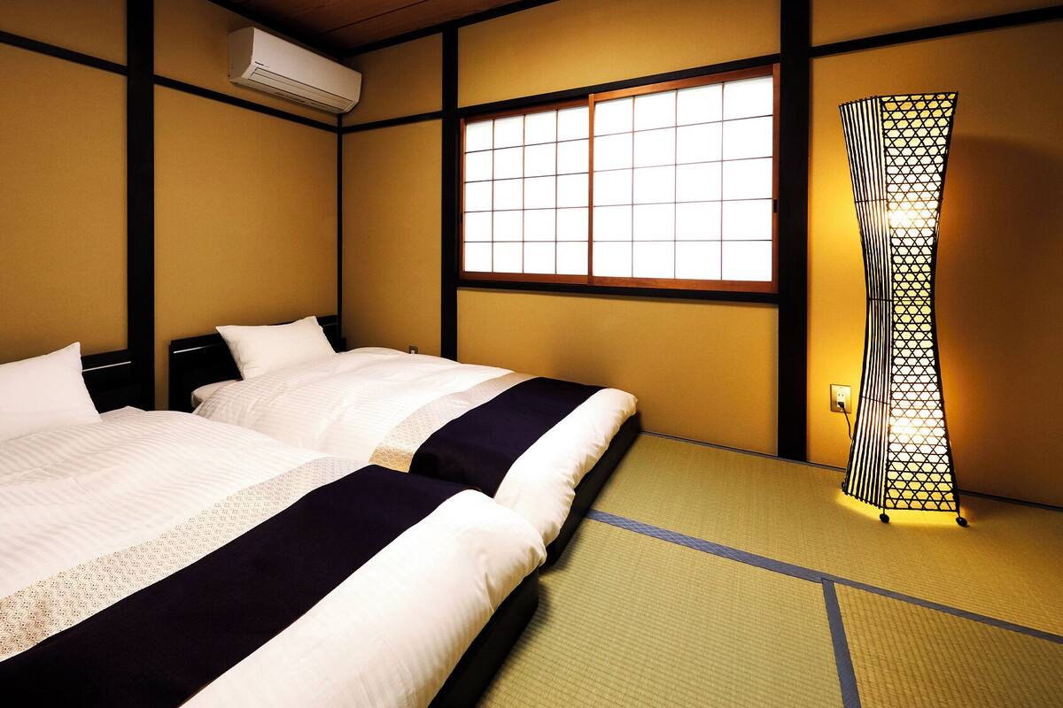 A traditional Japanese-style room with tatami flooring, two beds with white bedding, a sliding door with a geometric pattern, and a window with a grid design.