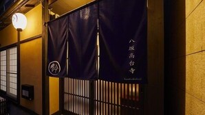 Fassade eines traditionellen japanischen Gebäudes mit einem dunkelblauen Eingangstor, einer kleinen beleuchteten Beschilderung und einem hölzernen Podest zum Eingang.