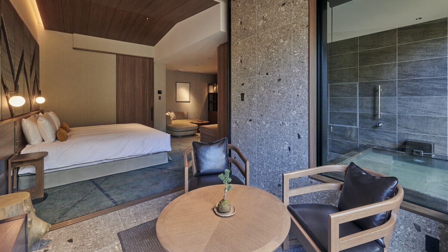 A modern hotel room with a bed, seating area, and a view of a stone wall and a glass shower.