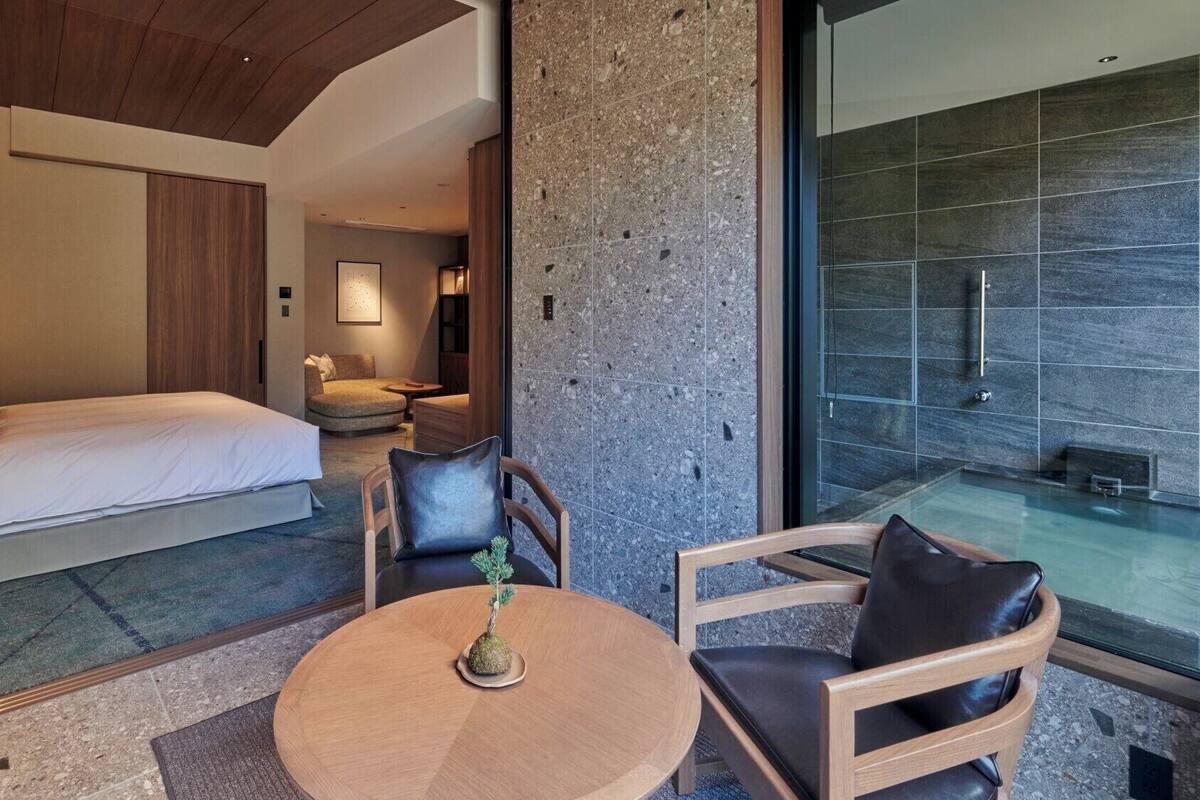 Quarto de hotel moderno com cama, área de estar e banheiro com paredes de vidro.