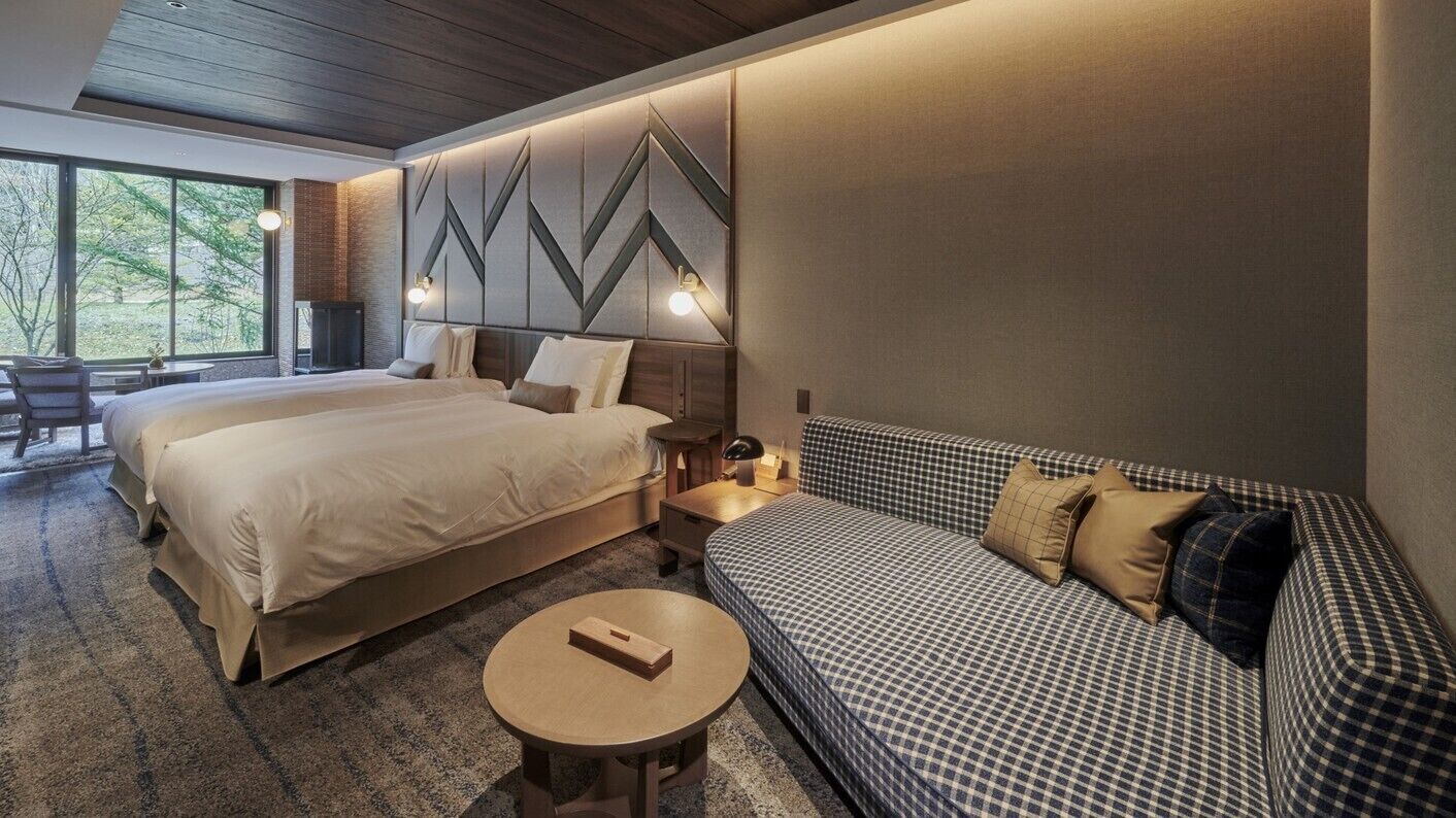 A hotel room with two beds, a sofa, a round table, and a view of trees through large windows.