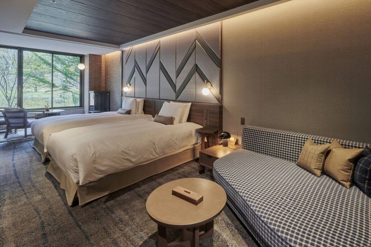 Quarto de hotel com duas camas, um sofá, uma mesa redonda e vista para árvores através de amplas janelas.
