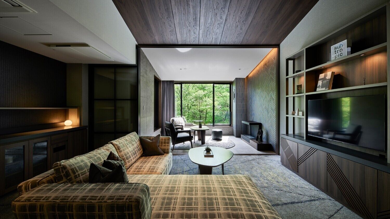 A modern living room with a sofa, coffee table, and TV. There is a large window with a view of greenery.