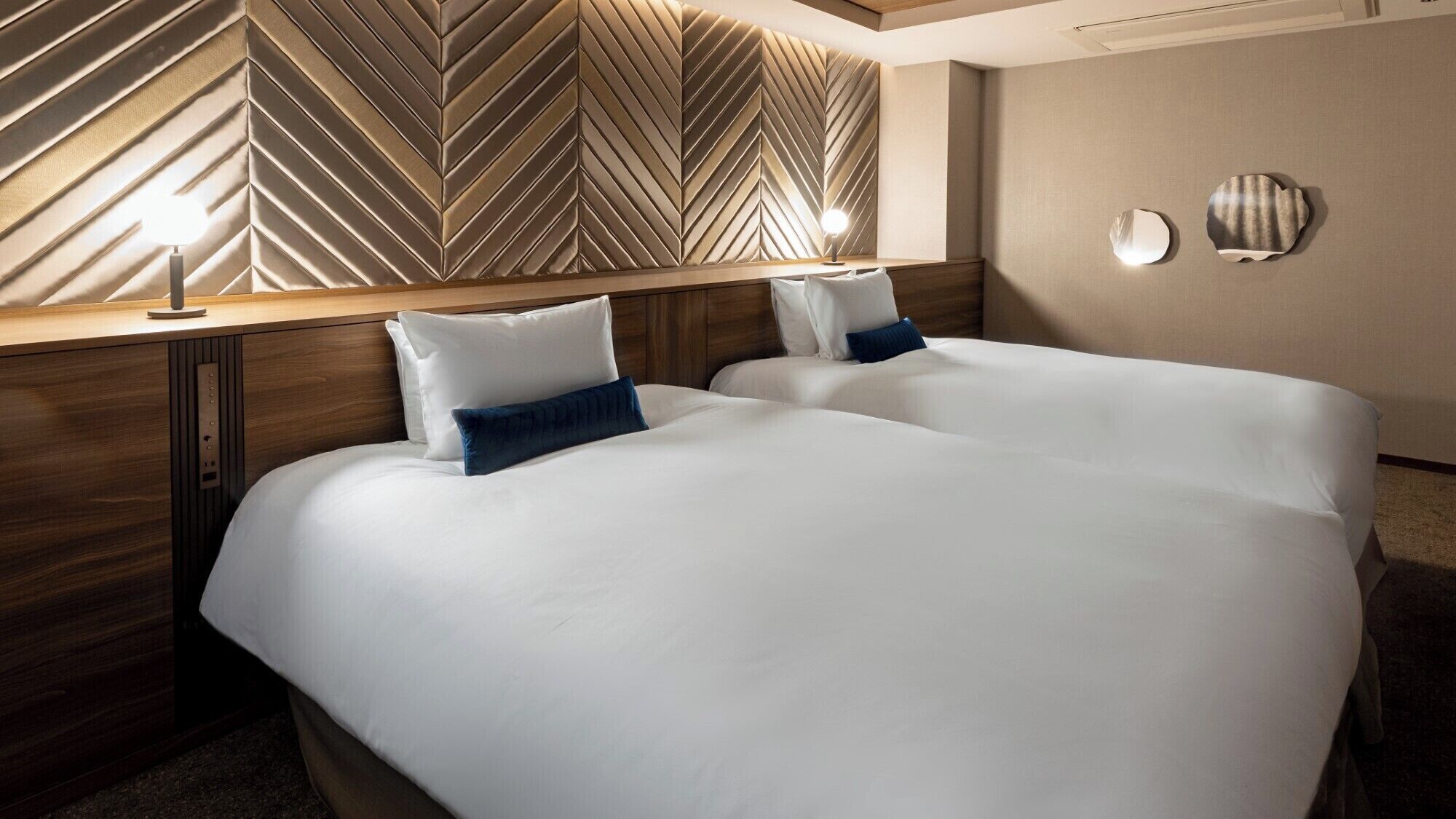 A hotel room with two beds, a wooden headboard with a chevron pattern, and wall-mounted lights.